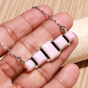 May include: A silver chain necklace featuring a horizontal bar pendant composed of five rectangular, light pink stones set in silver bezels. The stones are arranged in a graduated size pattern, with the largest stone positioned in the center.