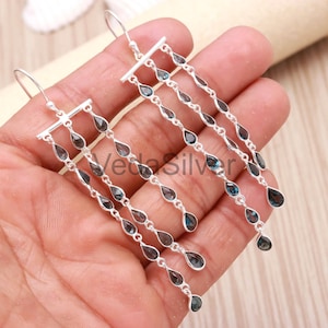 Può includere: Orecchini pendenti in argento con gemme blu scuro a forma di goccia. Gli orecchini hanno un design lineare con più fili di gemme sospese da una barra orizzontale. Le gemme sono collegate da piccoli anelli d'argento.