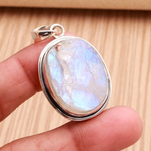 May include: An oval-shaped pendant featuring a polished moonstone with a translucent white appearance and iridescent blue flashes, set in a smooth silver metal frame with a small circular bail at the top for hanging.