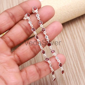 May include: Dangling silver earrings with a linear design. They feature a sequence of red gemstones and clear, smaller stones. The earrings are held in a hand, highlighting their elegant and elongated form.