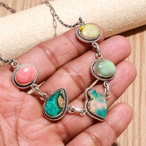 May include: A silver chain necklace featuring five distinct bezel-set gemstones in oval, teardrop, and spade shapes. The stones include a pink cabochon, a yellow-green stone, a light green stone, a turquoise stone, and a multi-colored opal-like piece.