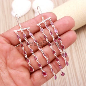 May include: Dangling silver earrings with multiple strands of teardrop-shaped, deep red gemstones. The earrings have a bar at the top with a hook for the ear. The gemstones are evenly spaced along the strands.