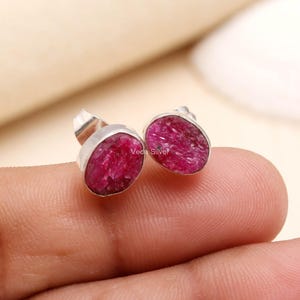 May include: A pair of stud earrings featuring raw, deep pink ruby stones set in polished silver metal. Each earring has an oval-shaped, textured gemstone surface with a metallic backing. The text Veda Silver is visible on the stone surface.
