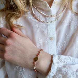 May include: A delicate bracelet with pink and white beads, gold accents, and a small gold disc. A layered pearl necklace is also visible. The jewelry is set against a white, embroidered blouse.