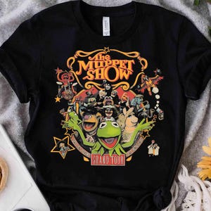 Puede incluir: Una camiseta negra de manga corta con un gráfico colorido de los personajes de The Muppet Show. El diseño incluye el texto The Muppet Show y Grand Tour, rodeado de varios Muppets, estrellas amarillas y adornos decorativos dorados.
