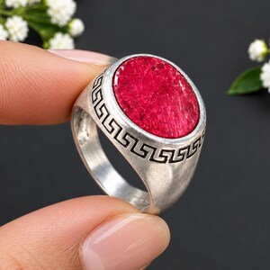 Peut inclure: Une bague argentée ornée d'une grande pierre rouge ovale à la texture marquée. L'anneau est décoré d'un motif grec gravé en noir qui entoure la pierre, créant un contraste saisissant avec la finition métallique argentée de la monture.