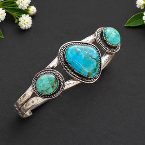 May include: A silver cuff bracelet featuring three turquoise stones in varying shapes and sizes. The central stone is large and irregular, flanked by two smaller, circular turquoise stones, all set in decorative silver bezels with rope detailing.