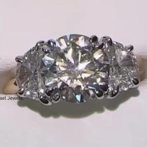 Puede incluir: Un anillo de tres piedras con un diamante central grande de talla brillante redonda flanqueado por dos diamantes laterales más pequeños, montado en una banda de metal pulido. El texto Lael Jewels es visible en el lado izquierdo.