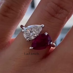 May include: A silver ring featuring two pear-shaped gemstones. One is a clear, sparkling diamond, and the other is a deep red ruby. The ring is set against a blurred background, with the text "Lael Jewels" visible.