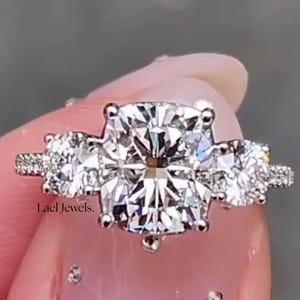 Puede incluir: Un anillo de compromiso de tres piedras con un diamante central grande de talla cuadrada y dos piedras laterales más pequeñas. El anillo está engastado en una banda plateada, con piedras de acento más pequeñas a lo largo de la banda.