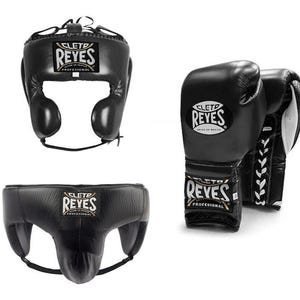 May include: Black boxing headgear, gloves, and groin protector set. The headgear has ear protection and a chin strap. The gloves are laced up and feature the brand name "REYES". The groin protector has a supportive design.