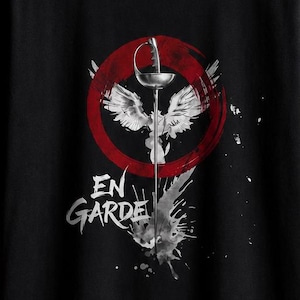 Puede incluir: Una camiseta negra con un estampado gráfico de un florete de esgrima plateado centrado dentro de un círculo rojo, acompañado de alas blancas y un efecto de salpicadura de tinta negra. El texto "EN GARDE" está impreso en letras blancas bajo la espada.