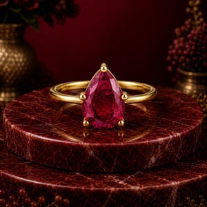 Puede incluir: Un anillo de oro con una piedra preciosa de color rojo intenso en forma de pera, sujeta por cuatro garras. La banda es lisa y pulida, mostrando un diseño sencillo que resalta el color vibrante y el corte facetado de la piedra central.