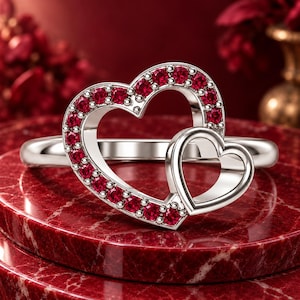 Puede incluir: Un anillo de tono plateado con dos formas de corazón entrelazadas. El corazón más grande está delineado con pequeñas piedras redondas de color rojo vibrante a lo largo de su borde, mientras que el más pequeño es de metal pulido.