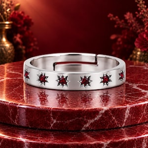 Puede incluir: Un anillo ajustable de tono plateado que presenta una serie de engastes en forma de estrella espaciados uniformemente. Cada estrella contiene una pequeña piedra preciosa redonda de color rojo intenso en el centro. El aro es liso y abierto.