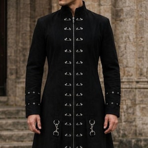May include: A long black coat with a high collar and decorative metal clasps. The coat features long sleeves and a flared skirt, creating a dramatic silhouette. The coat is made of a dark, possibly suede-like material, and is ankle-length.