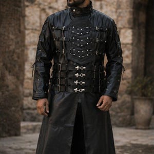 May include: A long black coat featuring a high collar and a front panel adorned with multiple horizontal straps, silver-toned buckles, and metallic studs. The garment has long sleeves and a floor-length hem, creating a structured appearance.