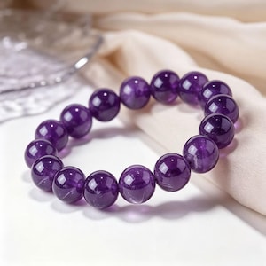 Pode incluir: Uma pulseira elástica circular composta por contas de ametista de cor roxa profunda, lisas e polidas. As contas são uniformes em tamanho e forma, apresentando uma superfície brilhante e reflexiva que destaca a translucidez natural da gema.
