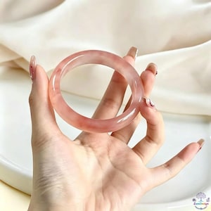 May include: A smooth, translucent bangle bracelet in a soft, pale pink hue with subtle, cloudy white marbling throughout the material. The circular accessory features a polished, glossy finish and a rounded profile, showcasing a consistent thickness.