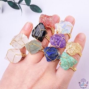 May include: A hand displays a collection of gemstone rings. Each ring features a raw, uncut stone in various colors, including pink, black, purple, and blue, secured by gold-colored wire. The rings are a unique jewelry item.