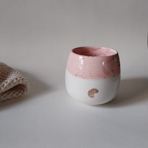 Könnte beinhalten: Ein Set aus drei Keramikbechern mit gesprenkelter, cremefarbener Glasur. Jeder Becher zeigt ein handgemaltes Design: ein blau-grünes Blatt, eine gelbe Blume und eine rote Beere. Die Becher haben eine einfache, abgerundete Form.