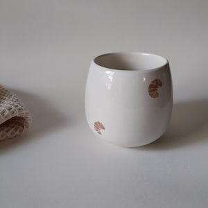 Könnte beinhalten: Eine weiße Keramik-Tasse mit schwarzem Henkel und dem Text "But First, Coffee" in fetter, schwarzer Schrift. Die Tasse steht vor einem verschwommenen Hintergrund, der eine gemütliche Umgebung suggeriert.
