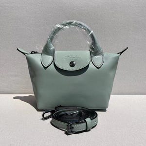May include: A light green handbag with a top handle and a detachable shoulder strap. The bag has a flap closure with a button and a logo. The strap is black with silver-colored hardware.