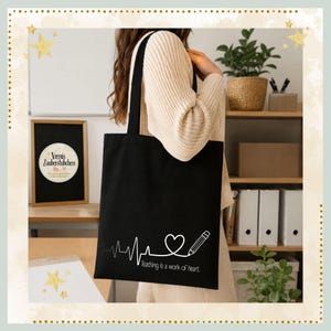 Könnte beinhalten: Schwarze Tragetasche mit weißer Grafik eines Herzschlags, eines Herzens und eines Bleistifts, mit dem Text "teaching is a work of heart". Die Tasche wird von einer Person in einem cremefarbenen Pullover getragen.