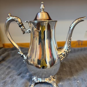 Könnte beinhalten: Eine weiße Keramik-Tasse mit schwarzem Henkel und dem Text "But First, Coffee" in fetter, schwarzer Schrift. Die Tasse steht vor einem verschwommenen Hintergrund, der eine gemütliche Umgebung suggeriert.