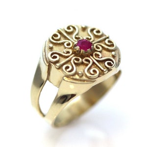 Może przedstawiać: Złoty pierścionek z ozdobnym, okrągłym wzorem. Pierścionek posiada centralny, okrągły, czerwony kamień szlachetny. Opaska ma podzielony wzór. Pierścionek jest umieszczony na białym tle.