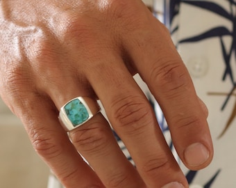 Könnte beinhalten: Ein silberner Ring mit einem quadratischen, türkisfarbenen Stein wird am Finger getragen. Ein silbernes Armband ist ebenfalls zu sehen. Die Person trägt ein weißes Hemd mit blauem und braunem Blättermuster.