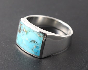 Könnte beinhalten: Ein silberner Ring mit einem rechteckigen Türkisstein. Der Ring hat ein poliertes, modernes Design. Der Türkisstein weist eine Mischung aus blauen und braunen Farben auf. Der Ring wird auf einer schwarzen Oberfläche präsentiert.