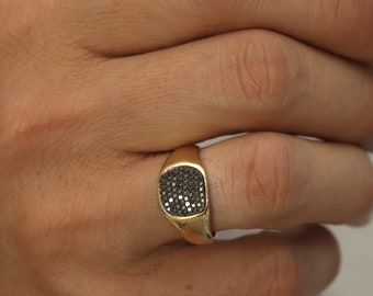 May include: A gold signet ring featuring a square-shaped face inlaid with numerous small, dark, reflective stones. The band is smooth and polished, with a tapered design that widens toward the setting, creating a classic and substantial appearance.