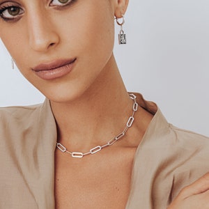 May include: A silver chain necklace and bracelet with a silver padlock charm earring. The jewelry is worn by a person with brown hair and dark eyes.