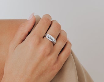 May include: A silver ring with the word "Believe" engraved on the top. The ring is worn on a person's finger.