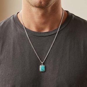 May include: A silver chain necklace with a rectangular turquoise pendant. The pendant is a vibrant blue-green colour. The necklace is worn over a grey t-shirt.