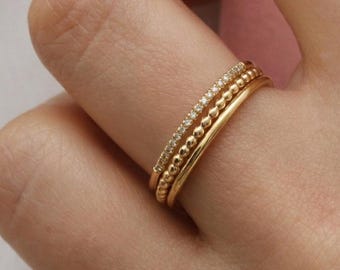 Può includere: Un set di tre anelli in oro indossati insieme su un dito. Il primo anello presenta una fascia sottile con piccole pietre chiare incastonate, il secondo ha un design a perline testurizzato e il terzo è una fascia in oro liscia.