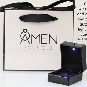 K&ouml;nnte beinhalten: Eine wei&szlig;e Geschenkt&uuml;te mit schwarzen Henkeln und dem Logo "AMEN BOUTIQUE". Vor der T&uuml;te befindet sich eine schwarze Ringbox mit einem Licht im Inneren. Der Text "You're welcome to add a unique ring box with automatic light to your purchase (See item details)" befindet sich auf der rechten Seite des Bildes.