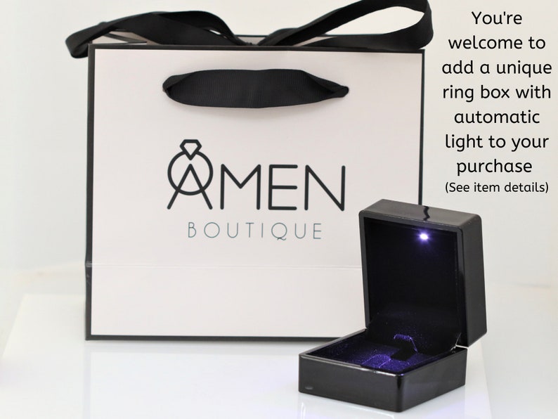 K&ouml;nnte beinhalten: Eine wei&szlig;e Geschenkt&uuml;te mit schwarzen Henkeln und dem Logo "AMEN BOUTIQUE". Vor der T&uuml;te befindet sich eine schwarze Ringbox mit einem Licht im Inneren. Der Text "You're welcome to add a unique ring box with automatic light to your purchase (See item details)" befindet sich auf der rechten Seite des Bildes.