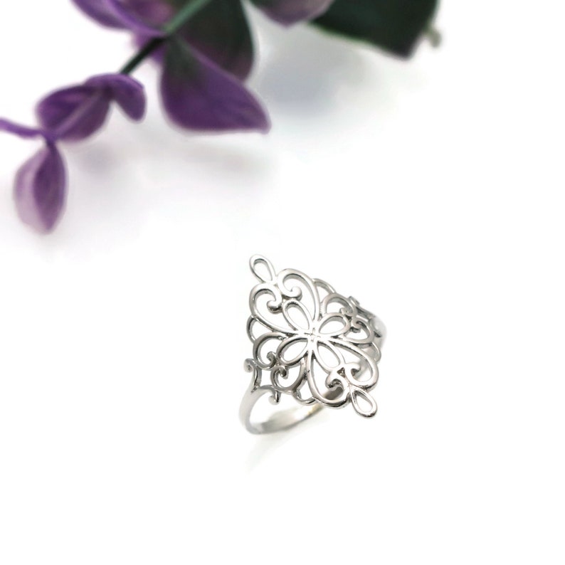 K&ouml;nnte beinhalten: Ein silberner Ring mit einem filigranen, komplizierten Design, das an eine Blume oder eine Schneeflocke erinnert. Der Ring besteht aus einem d&uuml;nnen Band und hat ein gro&szlig;es, durchbrochenes Design.