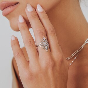 K&ouml;nnte beinhalten: Ein silberner Ring mit einem filigranen, komplizierten Design, das an eine Blume oder einen Stern erinnert. Der Ring wird am Zeigefinger einer Hand getragen.