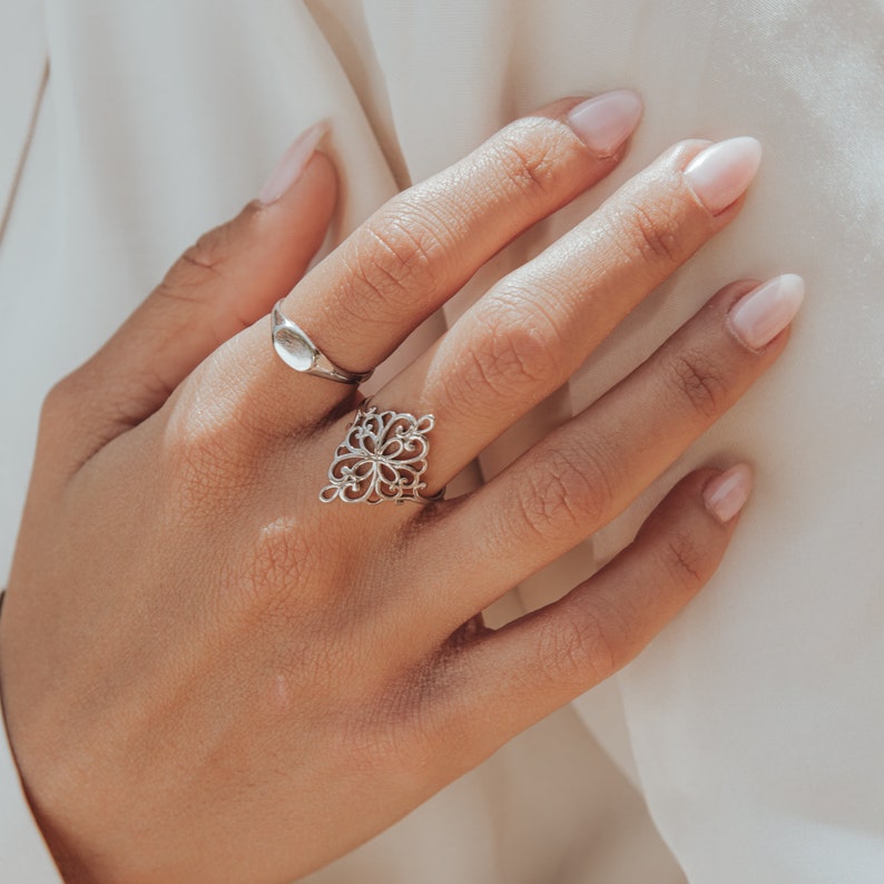 K&ouml;nnte beinhalten: Eine Hand tr&auml;gt zwei silberne Ringe. Der Ring am Zeigefinger ist ein filigranes Design. Der Ring am Mittelfinger ist ein schlichtes ovales Band.