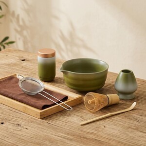 May include: A matcha tea set featuring a green ceramic bowl, whisk, and tea caddy. The set includes a bamboo scoop, a ceramic tea cup, a stainless steel strainer, and a wooden tray with a brown cloth.