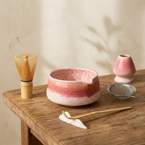 Peut inclure: Un ensemble à thé matcha sur une table en bois. L'ensemble comprend un bol en céramique rose et blanc, un fouet en bambou, une petite tasse à thé en céramique rose, une passoire en métal et une cuillère en bambou. Le bol a un bec verseur.