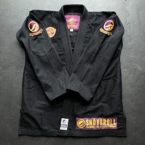 Può includere: Gi di Jiu-Jitsu nero con toppe ricamate viola e oro. Il gi presenta il marchio "SHOYOROLL" e altri loghi. Il gi è steso su una superficie grigia.
