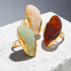 May include: Three statement rings with large, oval-shaped stones in cream, green, and brown hues. Each stone is set in a gold-colored, organic-shaped bezel and features a delicate gold band.