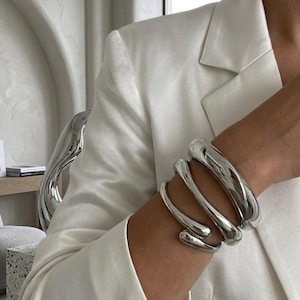 Op de afbeelding: Een zilverkleurige metalen armband met een meerlaags, spiraalvormig ontwerp met gladde, afgeronde randen. Het sieraad wikkelt zich in een continue, vloeiende beweging om de pols en heeft een gepolijst, reflecterend oppervlak dat het licht vangt.