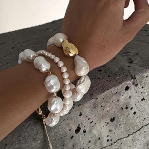 Könnte beinhalten: Eine Hand mit goldenen Ringen und Armbändern mit Perlen. Die Armbänder zeigen große, unregelmäßige weiße Perlen und ein goldenes Element. Ein Ring hat eine kleine Perle, der andere ist ein breites Goldband. Der Schmuck wird vor einem grauen Hintergrund präsentiert.