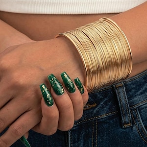 Peut inclure: Un large bracelet manchette doré composé de nombreux anneaux en fil métallique fin empilés. Le bracelet présente une finition métallique polie et une structure circulaire continue qui entoure le poignet, créant un aspect superposé et texturé.