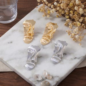 May include: Two pairs of earrings with an abstract, rippled, face-like design. One pair is finished in a polished gold tone, and the other pair is finished in a polished silver tone. Each earring features a smooth, undulating surface texture.